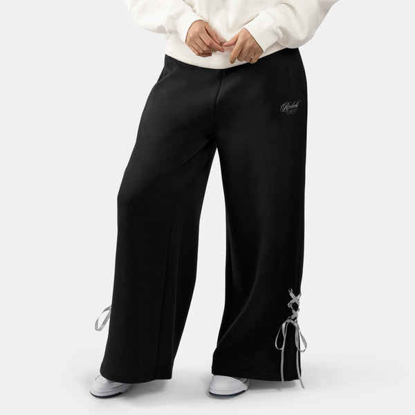 A person wearing black Glitz Ribbon Women's Jogger Pants with side lace-up details and a small script logo on the upper right thigh, paired with a white sweatshirt and white sneakers against a light gray studio background.